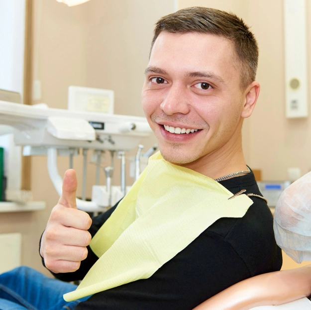 Young man giving thumbs up in orthodontics office, wearing yellow and black shirt, smiling at camera.