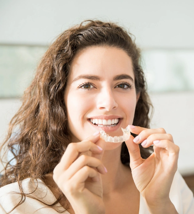 Woman holding clear aligner tray, smiling in bright clinical setting for orthodontics practice.