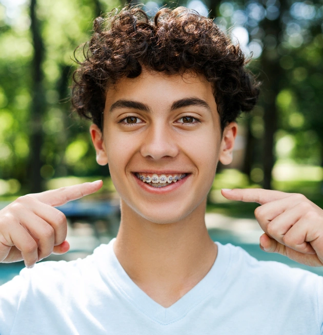 Smiling teenage boy pointing to teeth outdoors in casual white shirt, green background.